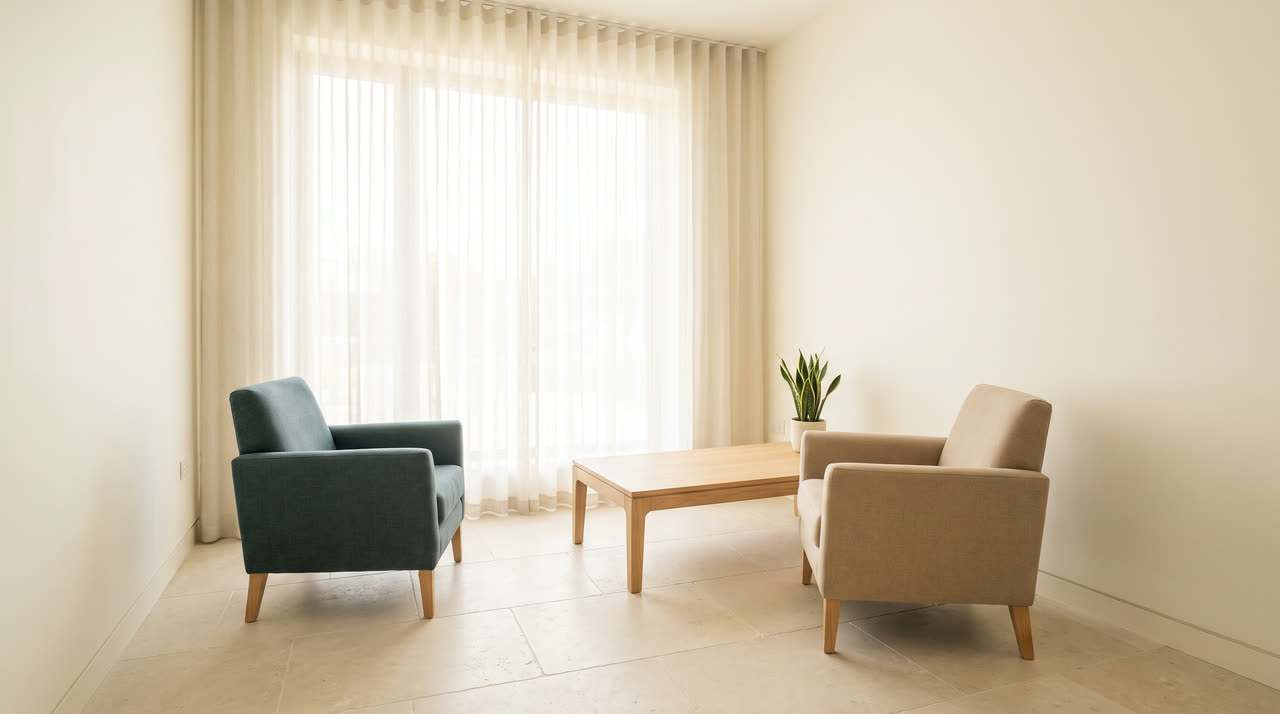 An empty, bright consultation room with two chairs and a low table by a large window.